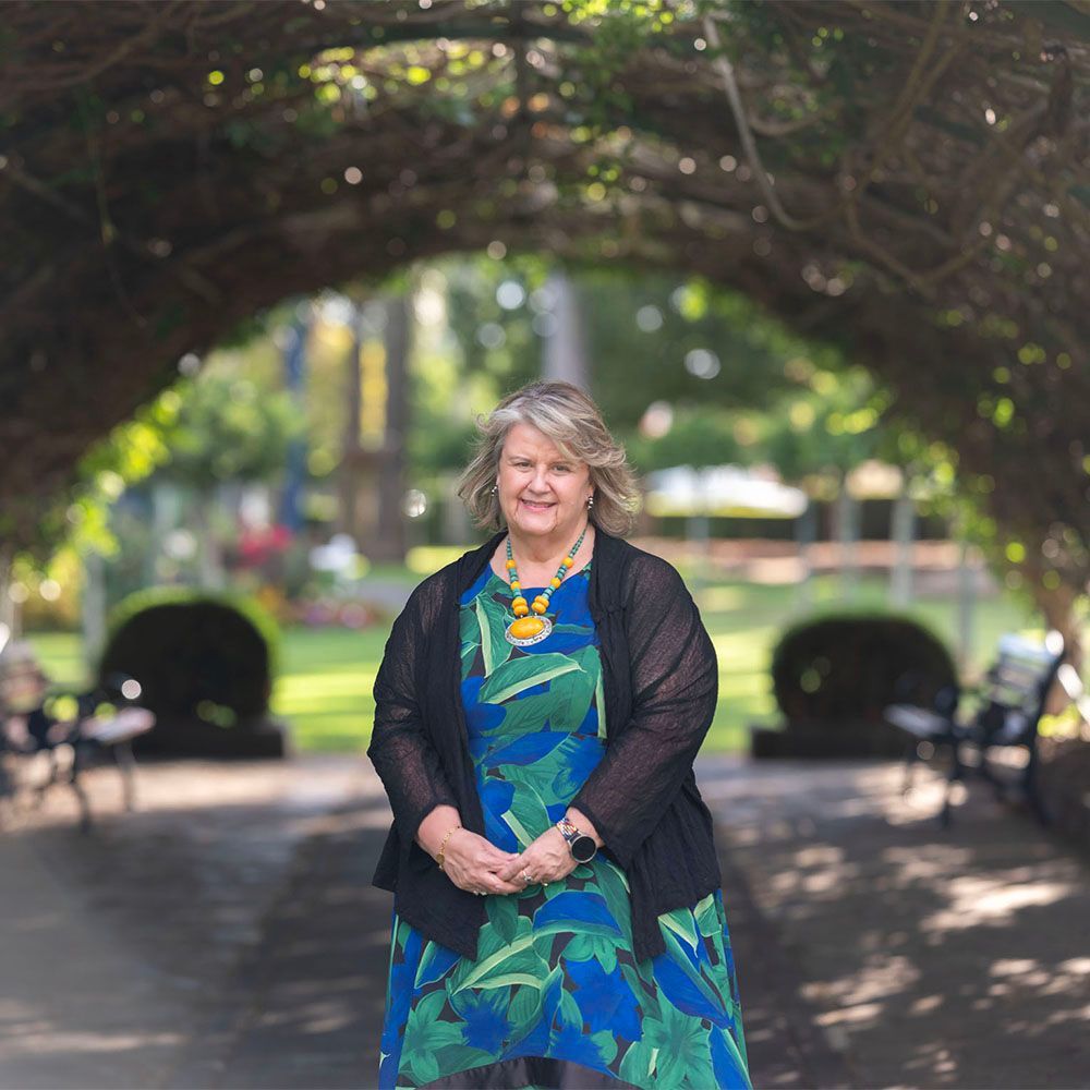 Woman in a blue-green dress standing under an arbor in a park, with trees and benches in the background.