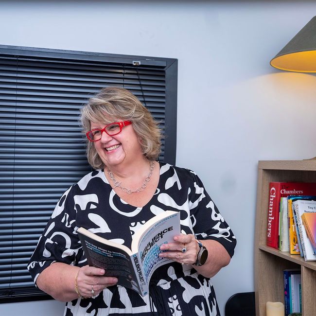 Smiling woman with red glasses reading a book beside a bookshelf in a cozy room