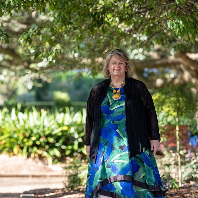 Woman in a blue-green dress and black shawl standing outdoors under a leafy tree