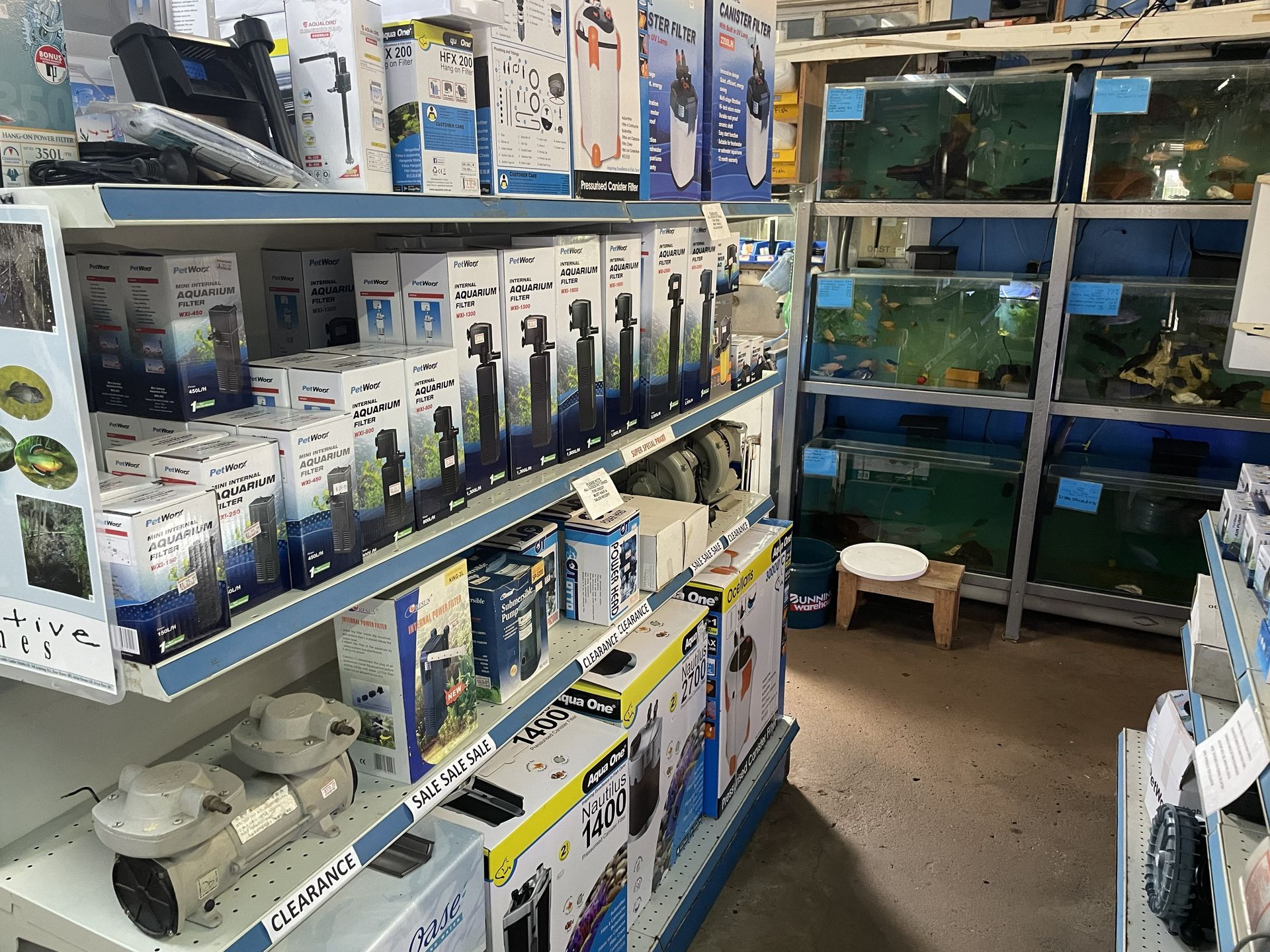 Shelves stocked with aquarium supplies in a pet store, tanks with fish in the background.