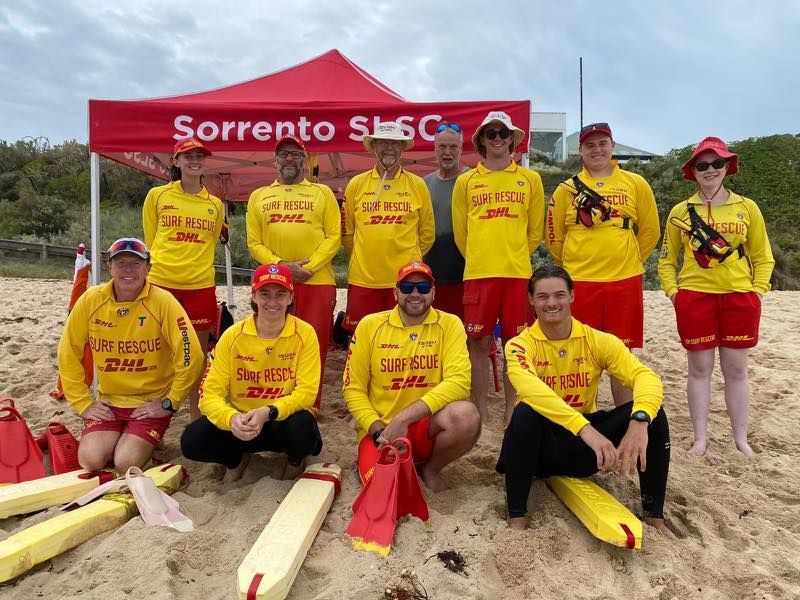 Group of lifeguards in yellow shirts and red shorts posing on a beach under a red canopy.