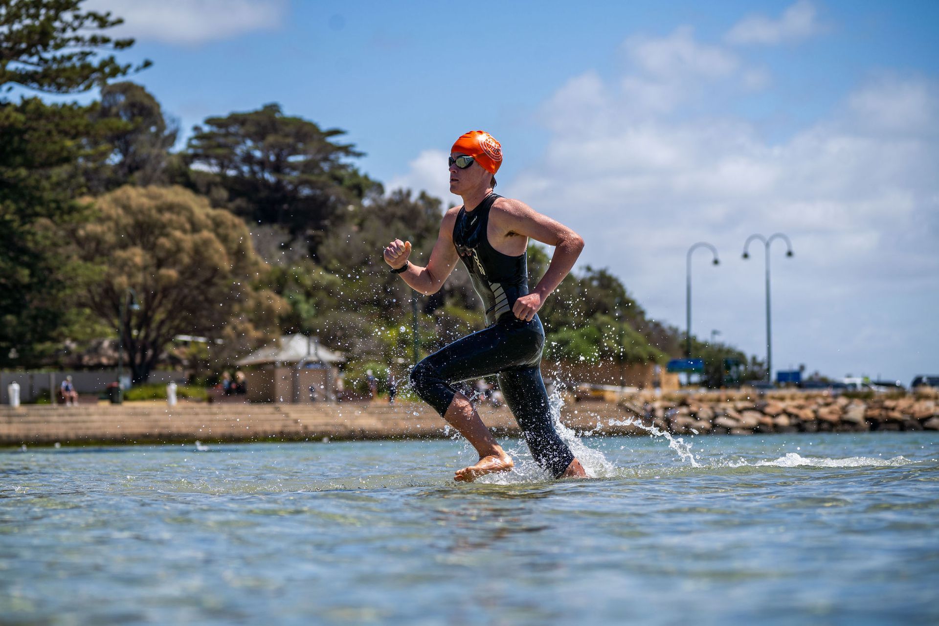 Triathletes running into ocean, waves crashing on shore. Blue sky, sunny day.