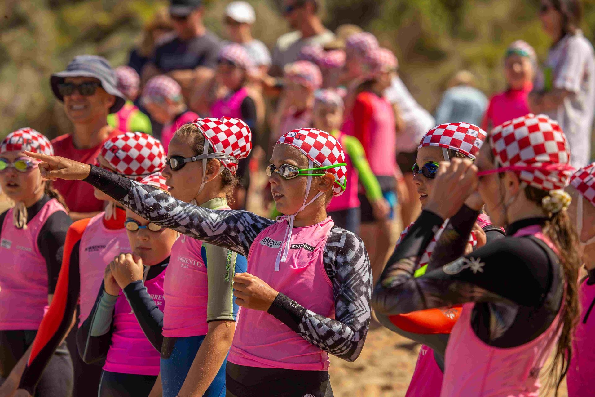 Young swimmers in pink, checkered caps and goggles, listening to instructions on a beach.