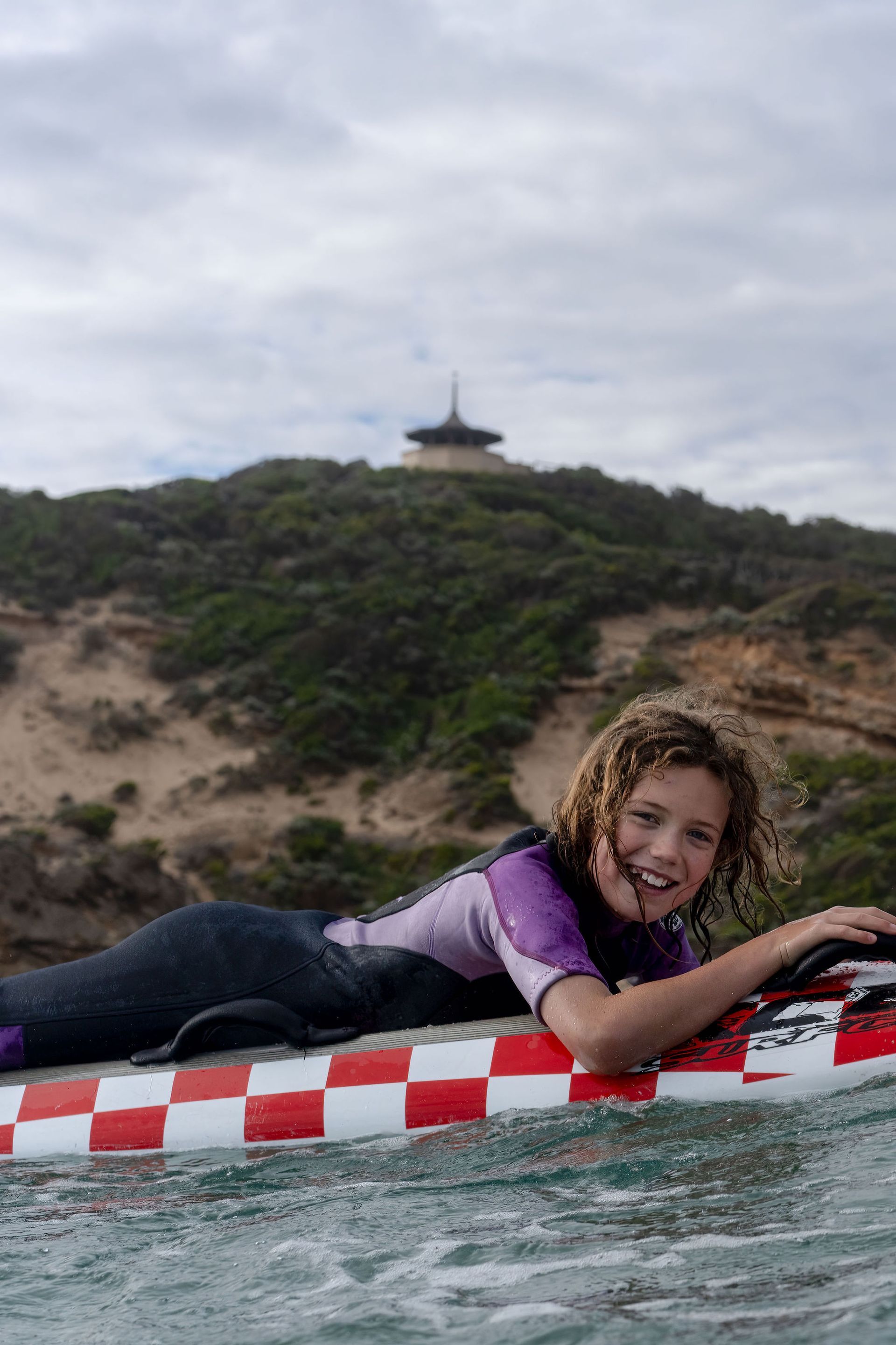 Girl on surfboard smiles, water, coastal hill, structure in background, wearing wetsuit.