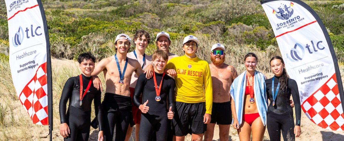 Club Championship competitors on the beach with medals, in front of TLC Healthcare and Sorrento SLSC flags.