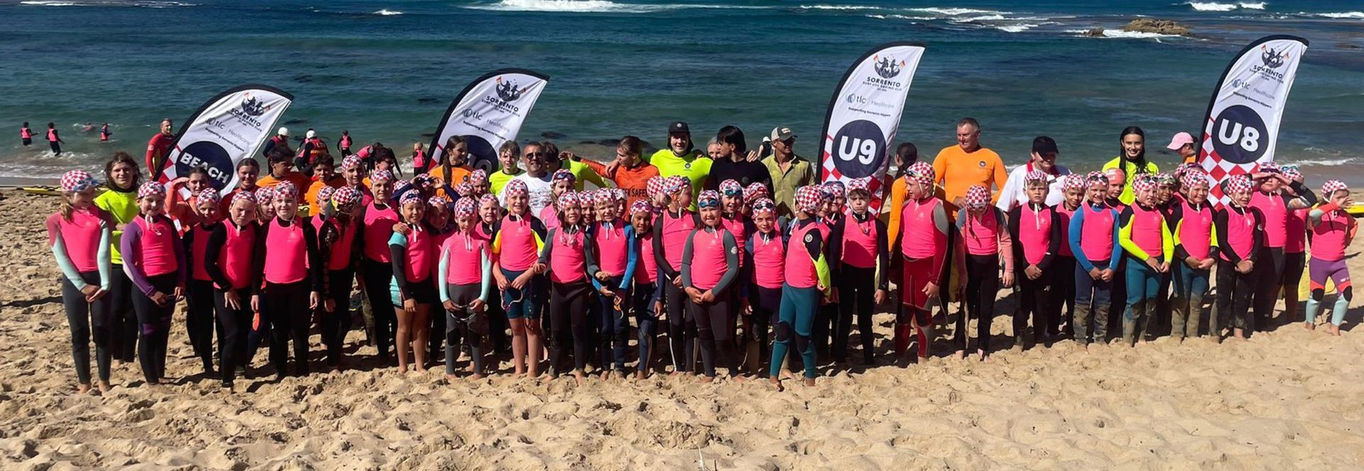 The full Sorrento nippers cohort lined up on the sand in pink and yellow rash vests behind age group flags.