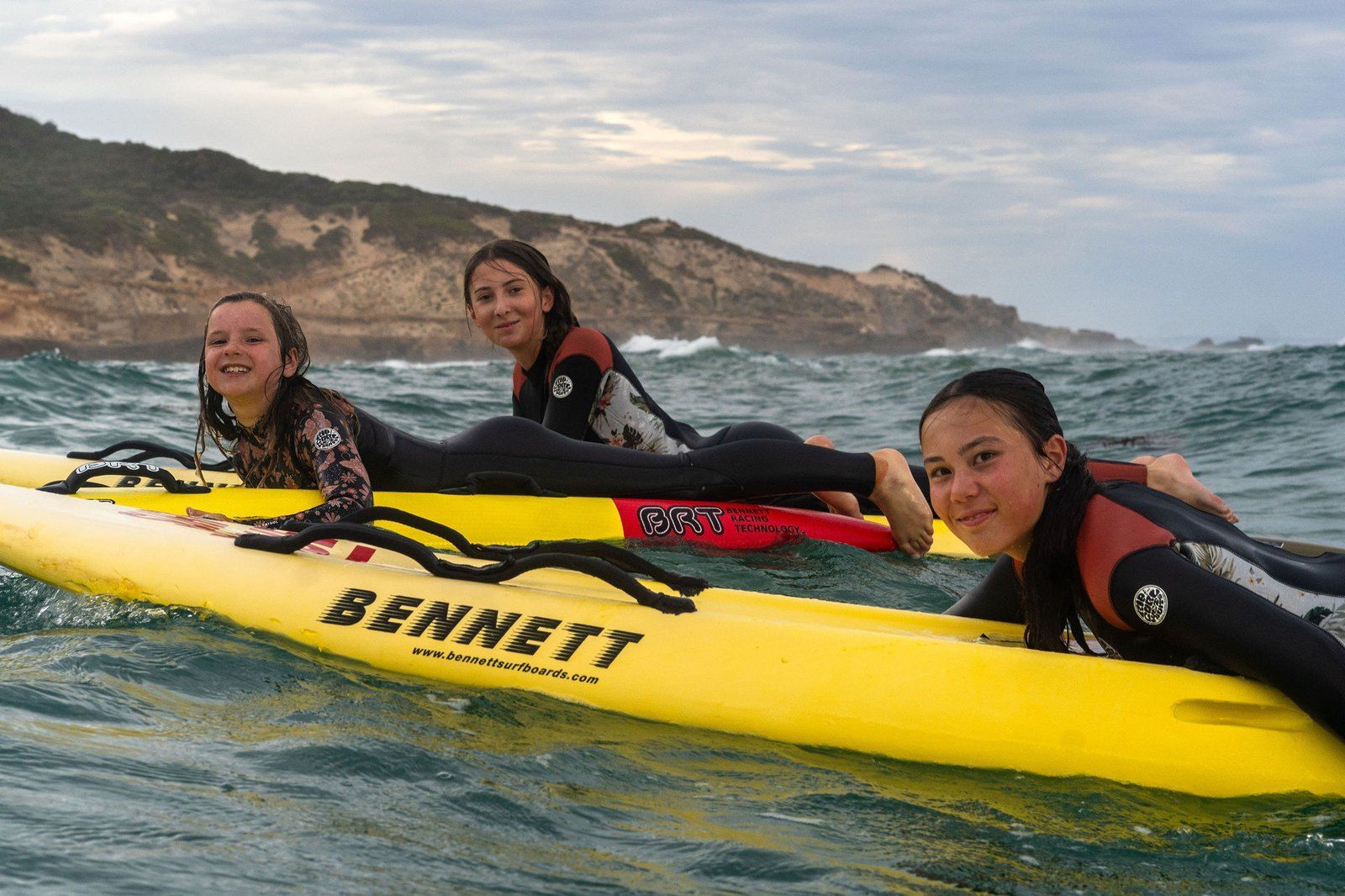 Three young members resting on a rescue board in the water at a Thursday night paddle out.