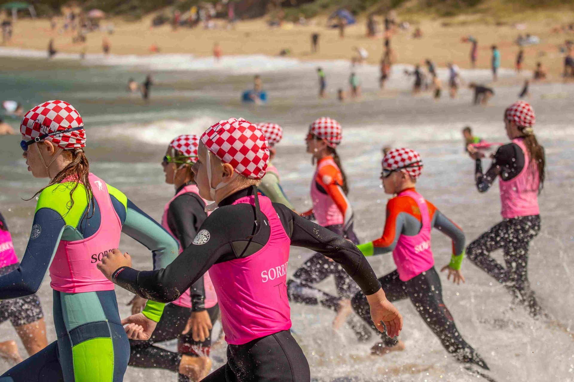 Youth running out of ocean, wearing pink, black and neon wetsuits and checkered swim caps.