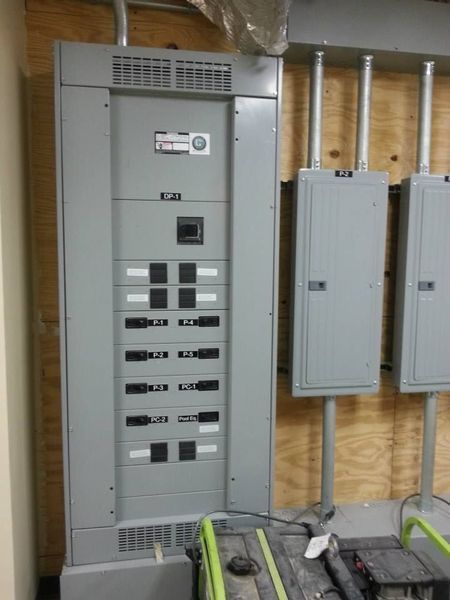 Gray electrical panel with multiple breakers and conduit pipes on a wooden wall.