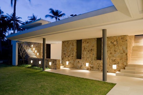 Modern house exterior with stone walls, pillars, and outdoor lighting. Grass and walkway lead to the entrance.