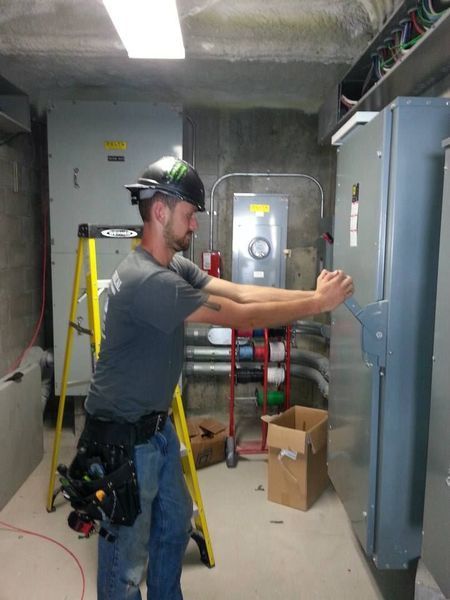 Electrician in a gray shirt and hard hat opening an electrical panel in a utility room.