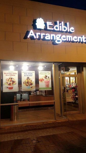 Edible Arrangements storefront, nighttime view. Lit-up sign and window displays of fruit arrangements.