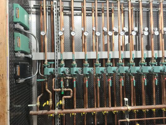 Copper pipes with valves and gauges, mounted on a wall. Green boxes and a dark electrical panel.