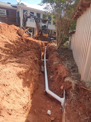 Gloved worker fitting a black drainage pipe into a trench with rocks and red soil