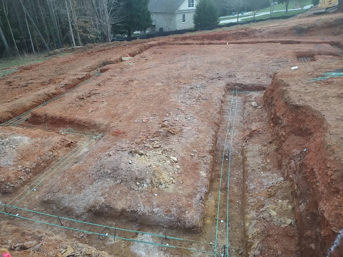 Cleared red dirt lot with trenches and a small house in the background