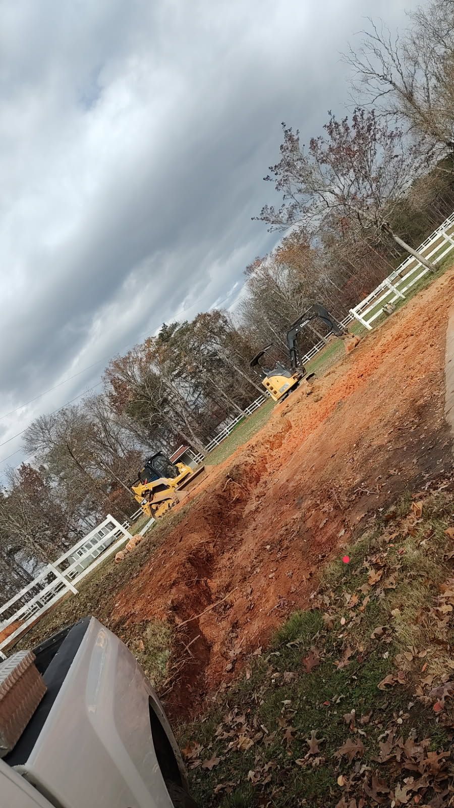 Tilted view of a rural yard with bare trees, white fence, and a cloudy sky over brown grass and leaves