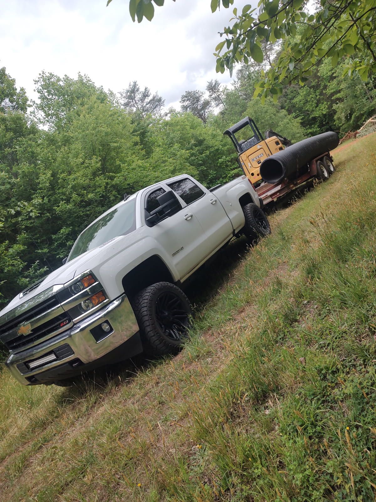 White pickup truck parked on a grassy slope, towing a black trailer in a wooded area.