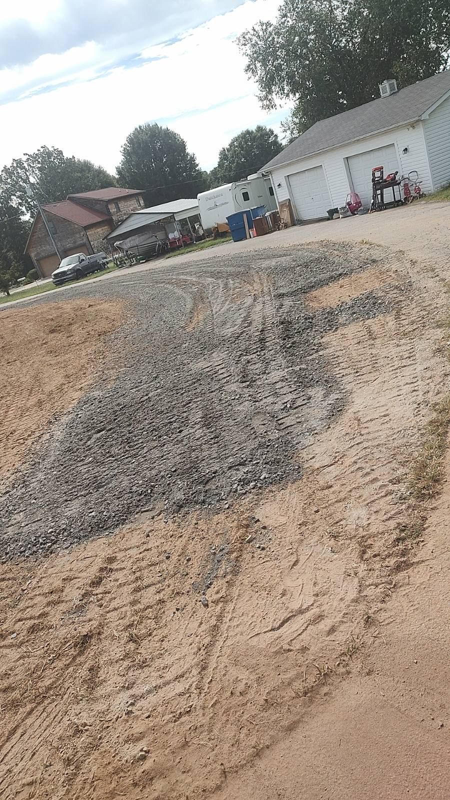 Gravelly dirt road beside white buildings and trees under a cloudy sky
