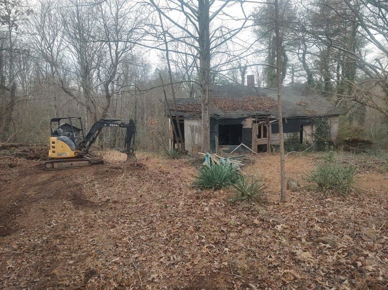 Excavator beside an abandoned house in a wooded, leaf-covered area