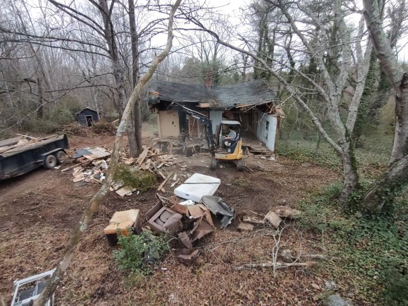 Dilapidated shed in a wooded yard, with debris and an old truck nearby