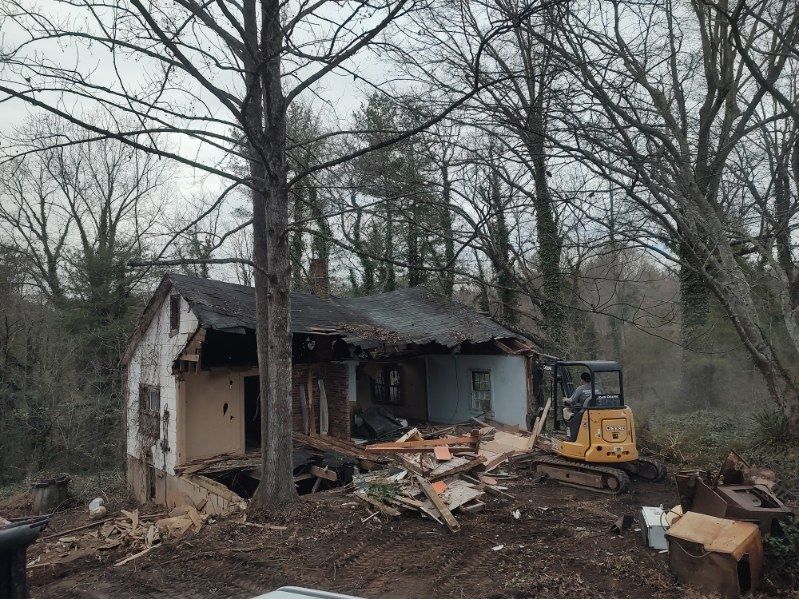 Partially demolished house with debris and a yellow skid steer in a wooded yard