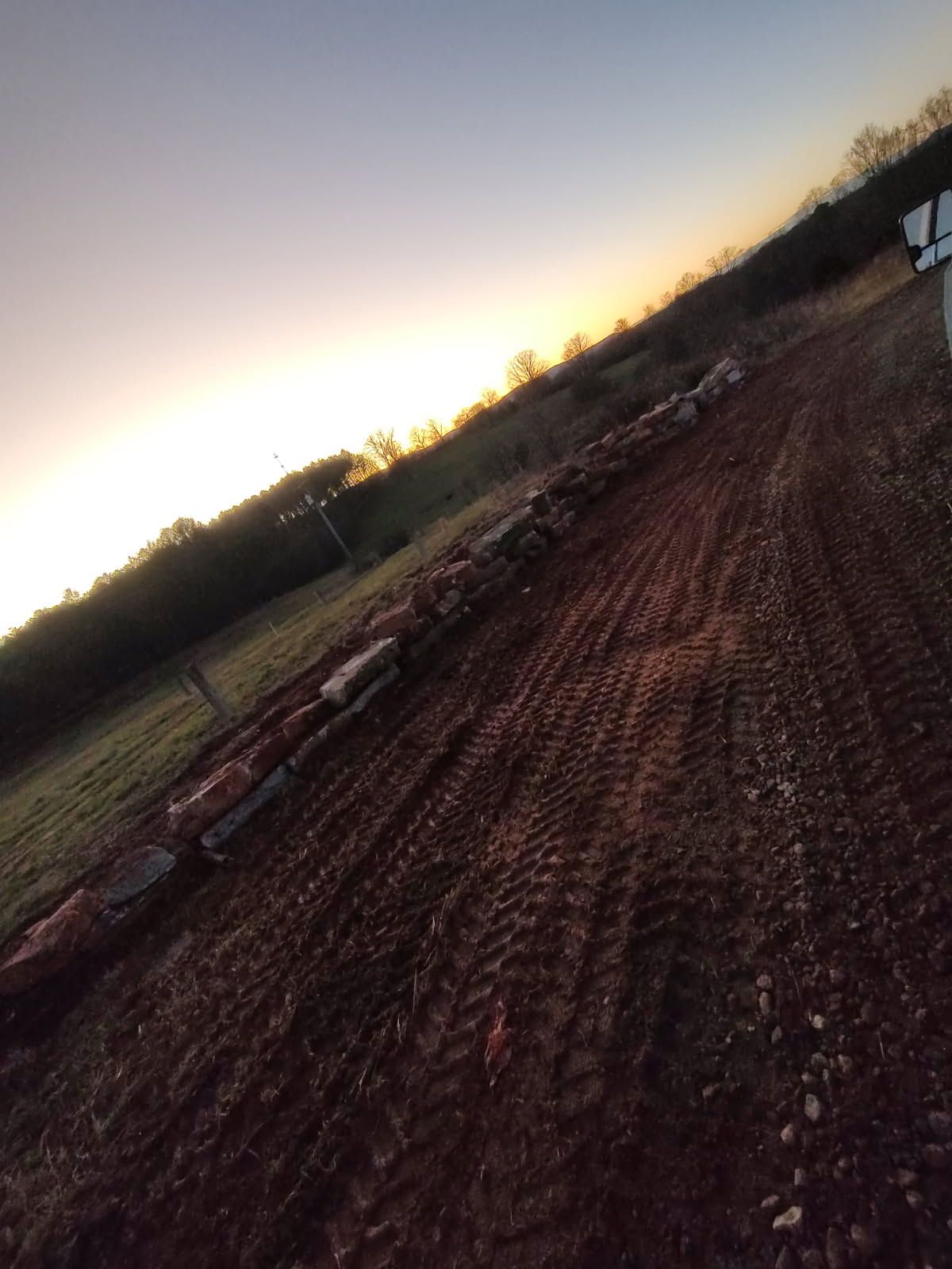 Tilted view of a freshly plowed field at sunset, with a dirt path and tree line on the horizon
