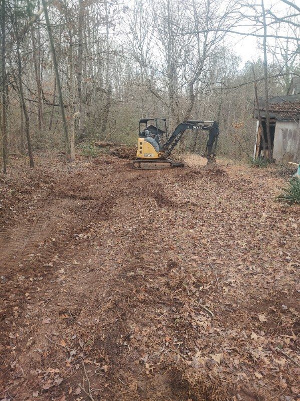 Small excavator clearing a leaf-covered dirt path in a leafless wooded area near a shed.