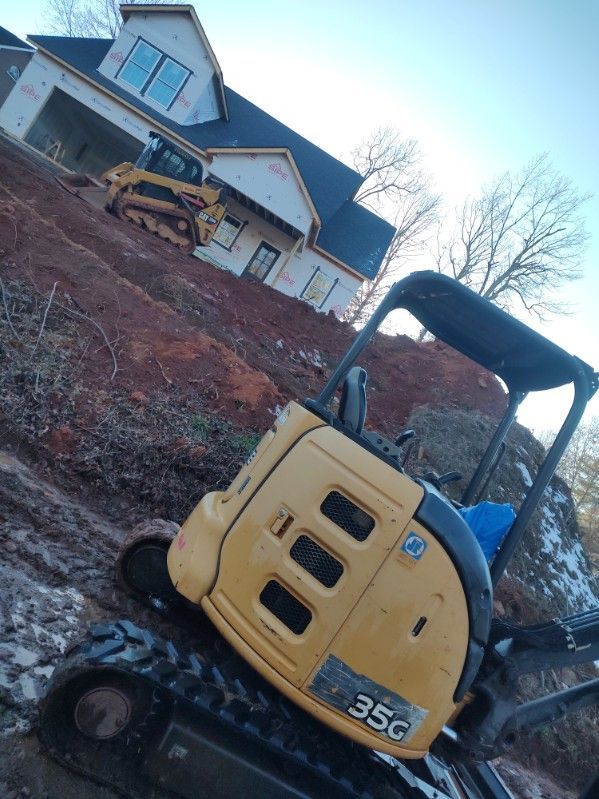 Yellow mini excavator on a sloped dirt lot beside a house under construction.
