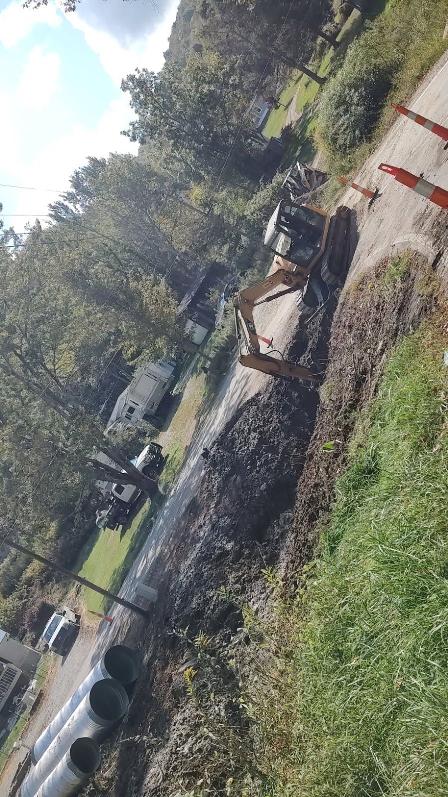 Roadwork scene with an excavator digging beside a damaged curb and a parked car on a sunny street