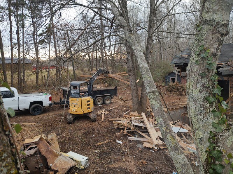Small yellow skid steer and white pickup in a wooded work site with cut trees and scattered debris