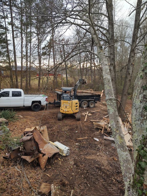 White pickup and yellow excavator in a wooded yard with cut tree stumps and fallen branches