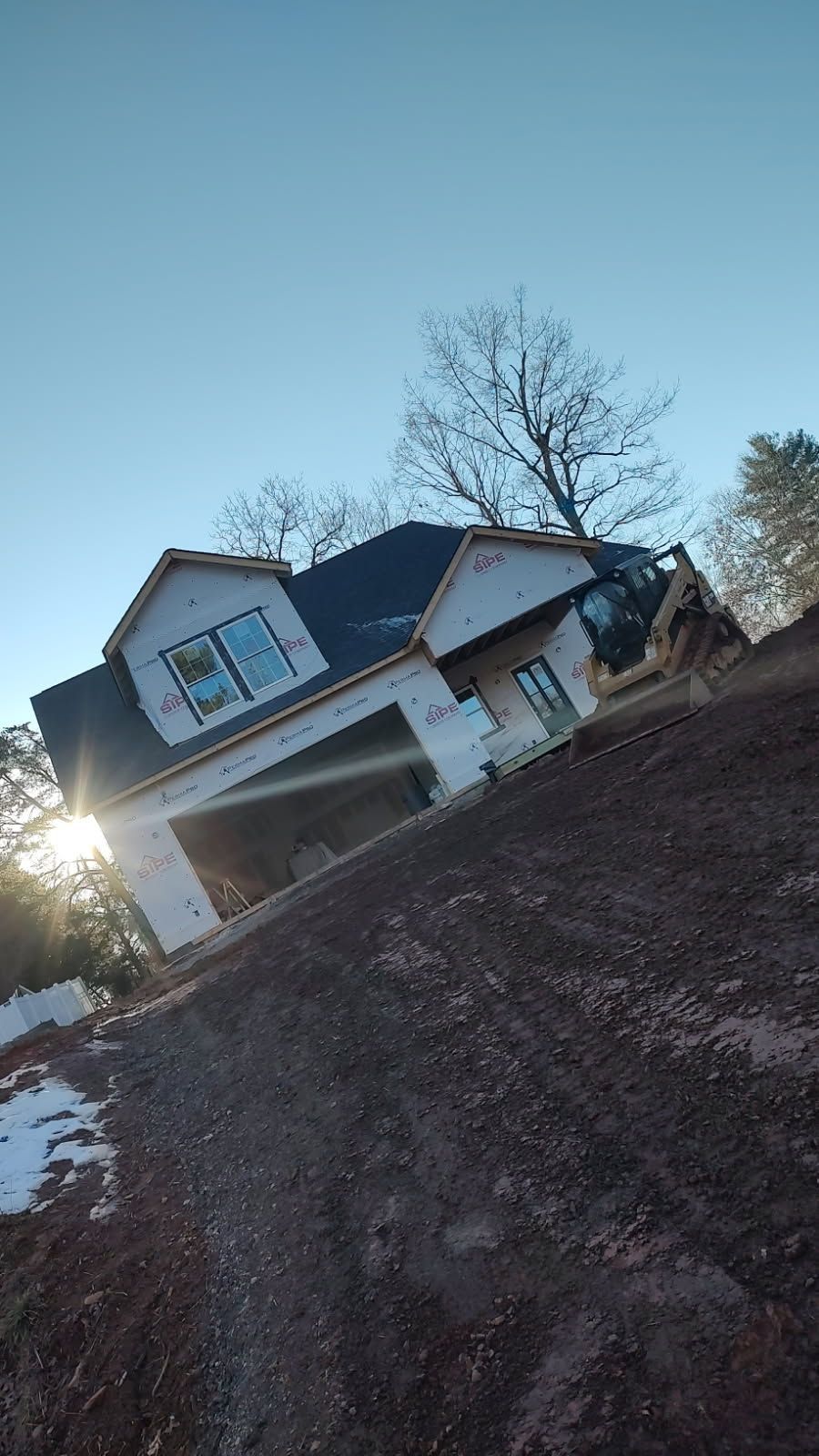 Tilted view of a suburban house with garage at sunset, beside a muddy dirt driveway and bare trees.