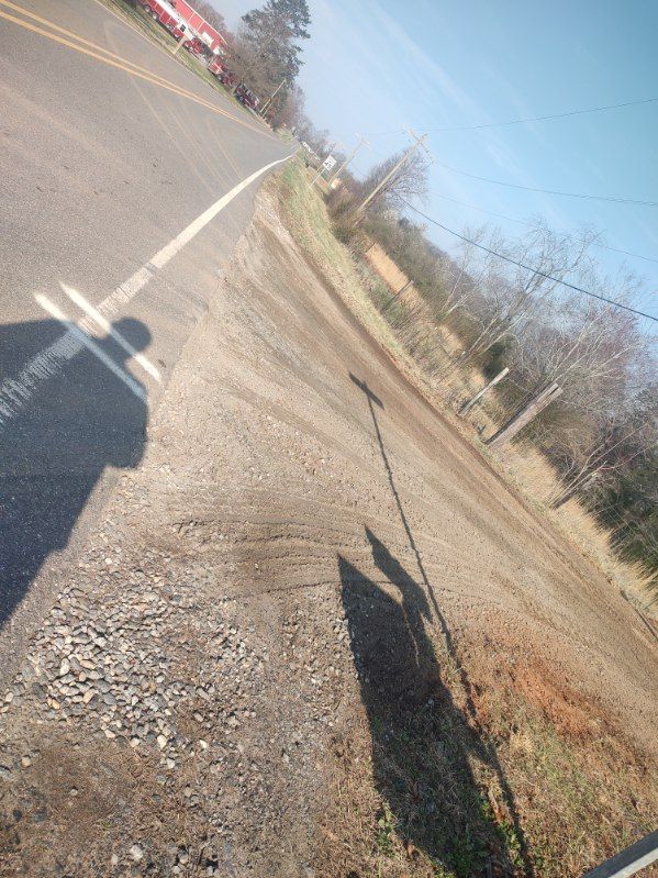 Tilted roadside view of a rural gravel shoulder beside a paved road, with bare trees and clear blue sky