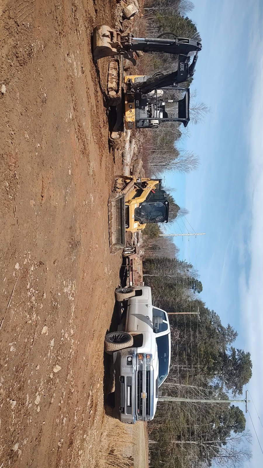 Construction site with excavator, dump trucks, and a white pickup on a dirt lot under a blue sky