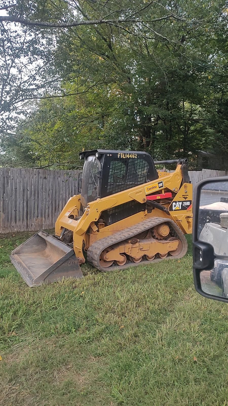 Yellow tracked skid steer on a grassy slope beside a wooden fence and trees.