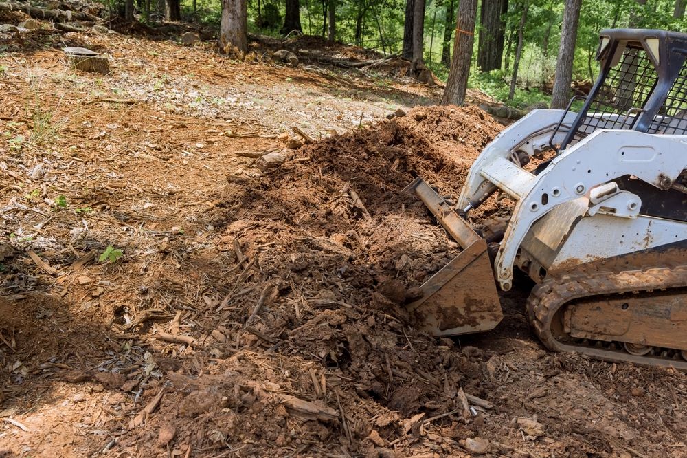 White skid-steer bulldozing dirt on a wooded trail, pushing a mound of soil