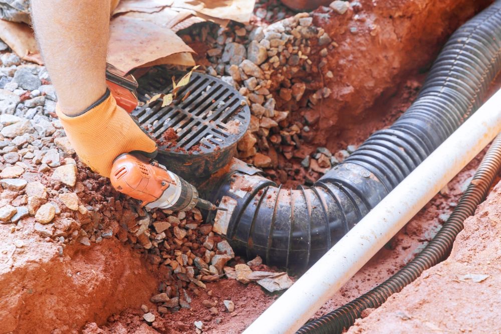 Orange-gloved worker fitting black drainage pipe in a dirt trench beside rocks and a white pipe