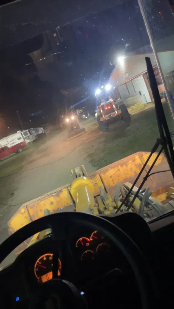 Cab view of a nighttime construction site, with machinery, bright work lights, and a road ahead.