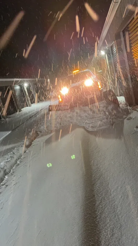Snowy night on a sloped street, with a bright car approaching and heavy snow falling.