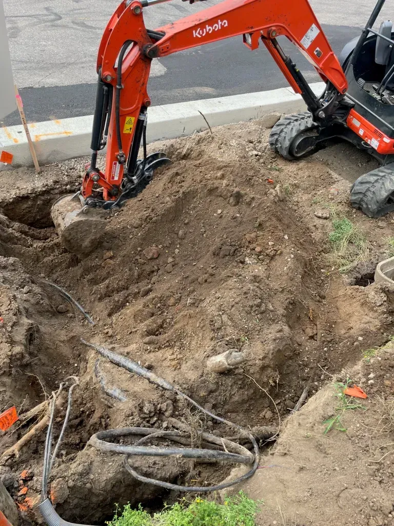 Orange excavator digging a trench in a dirt roadside area with exposed roots and debris
