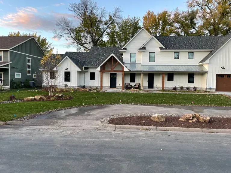 Suburban two-story house with white siding, dark roof, front porch, and driveway under autumn trees.