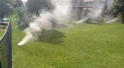 Sprinklers spraying white mist over a green lawn beside a chain-link fence and houses in the background
