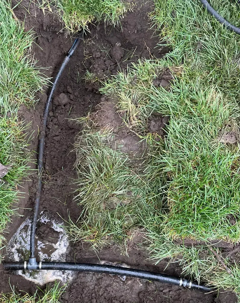 Black irrigation hose in a grassy dirt trench with muddy soil and a water leak near the bottom left.