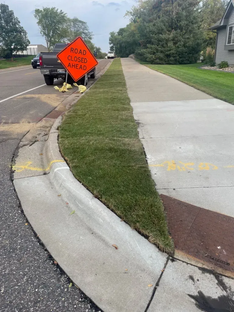 Broken curb and sidewalk by a roadwork sign near a driveway, with grass strip between concrete paths.