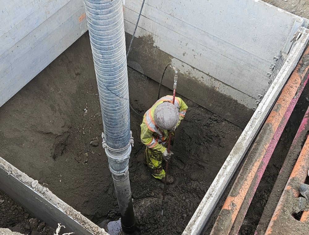 Worker in fluorescent safety gear standing in a concrete pit beside a large pipe.