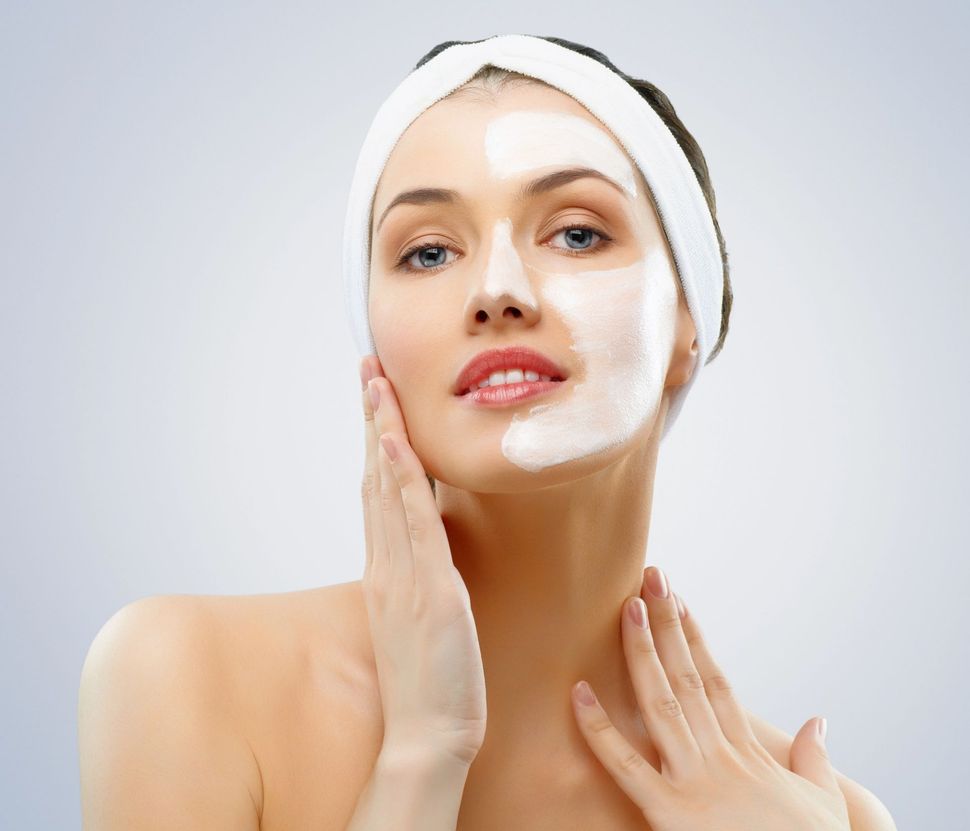 Woman with white towel headwrap applying skincare cream to her face against a light background