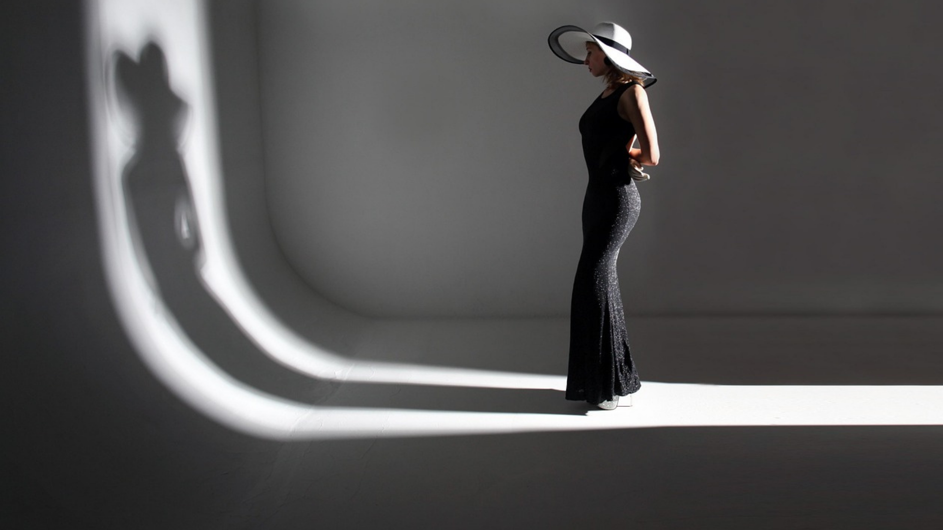 Une femme en robe et chapeau noirs se tient debout dans une pièce grise minimaliste, baignée d'un éclairage d'ombres.