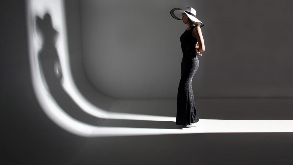 Une femme en robe et chapeau noirs se tient debout dans une pièce grise minimaliste, baignée d'un éclairage d'ombres.