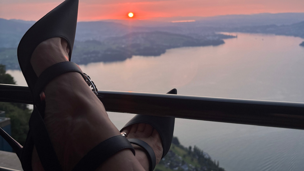 Les pieds détendus sur une rambarde surplombant un lac illuminé par le coucher du soleil et des montagnes lointaines