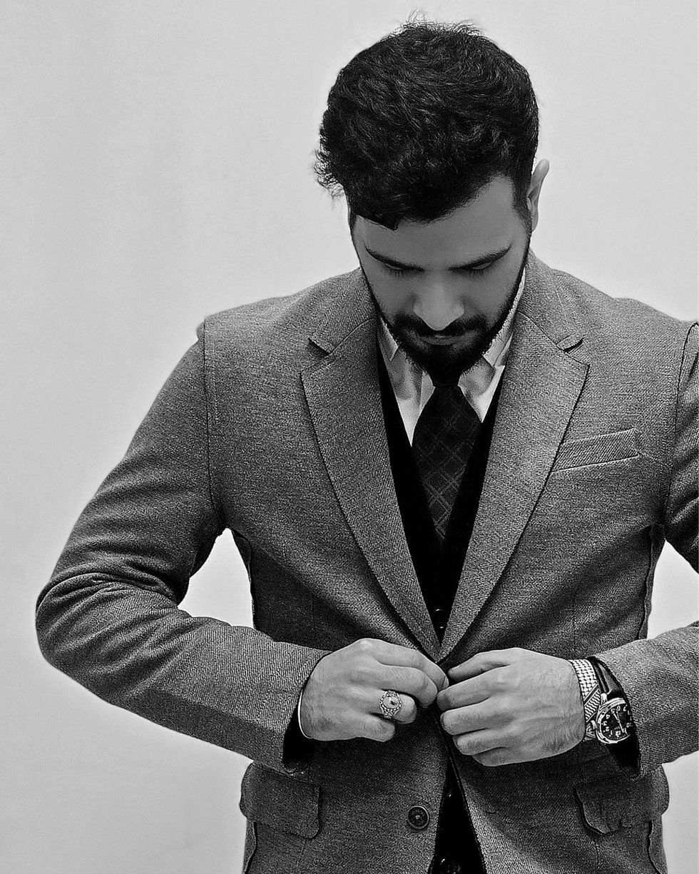 Un homme ajuste les boutons d'un blazer gris par-dessus une chemise et une cravate, le regard baissé.